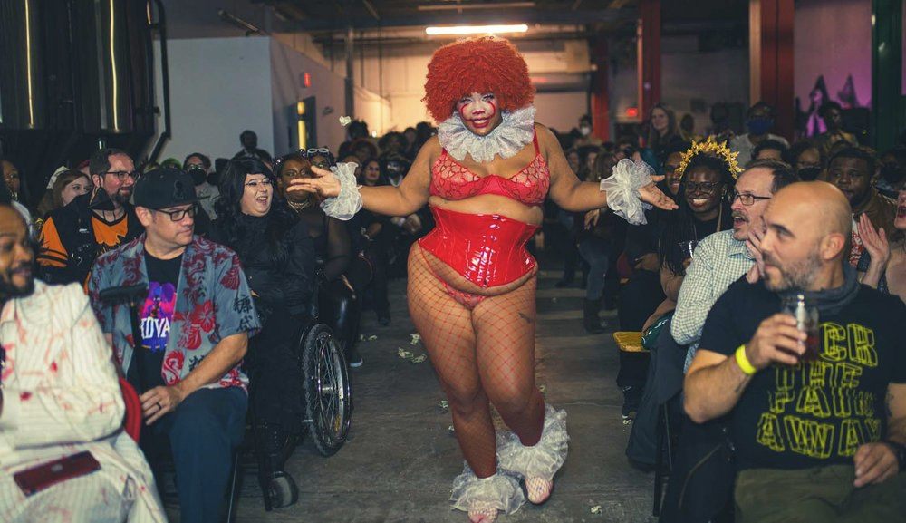 Photo of Rosie Beret walking down the aisle (she/they/he) dressed as a sexy version of IT. Rosie is wearing red lingerie and fishnet tights with a red waist cinching corset, white clown ruffle collar and matching ankle ruffles, with bright orange afro hair.